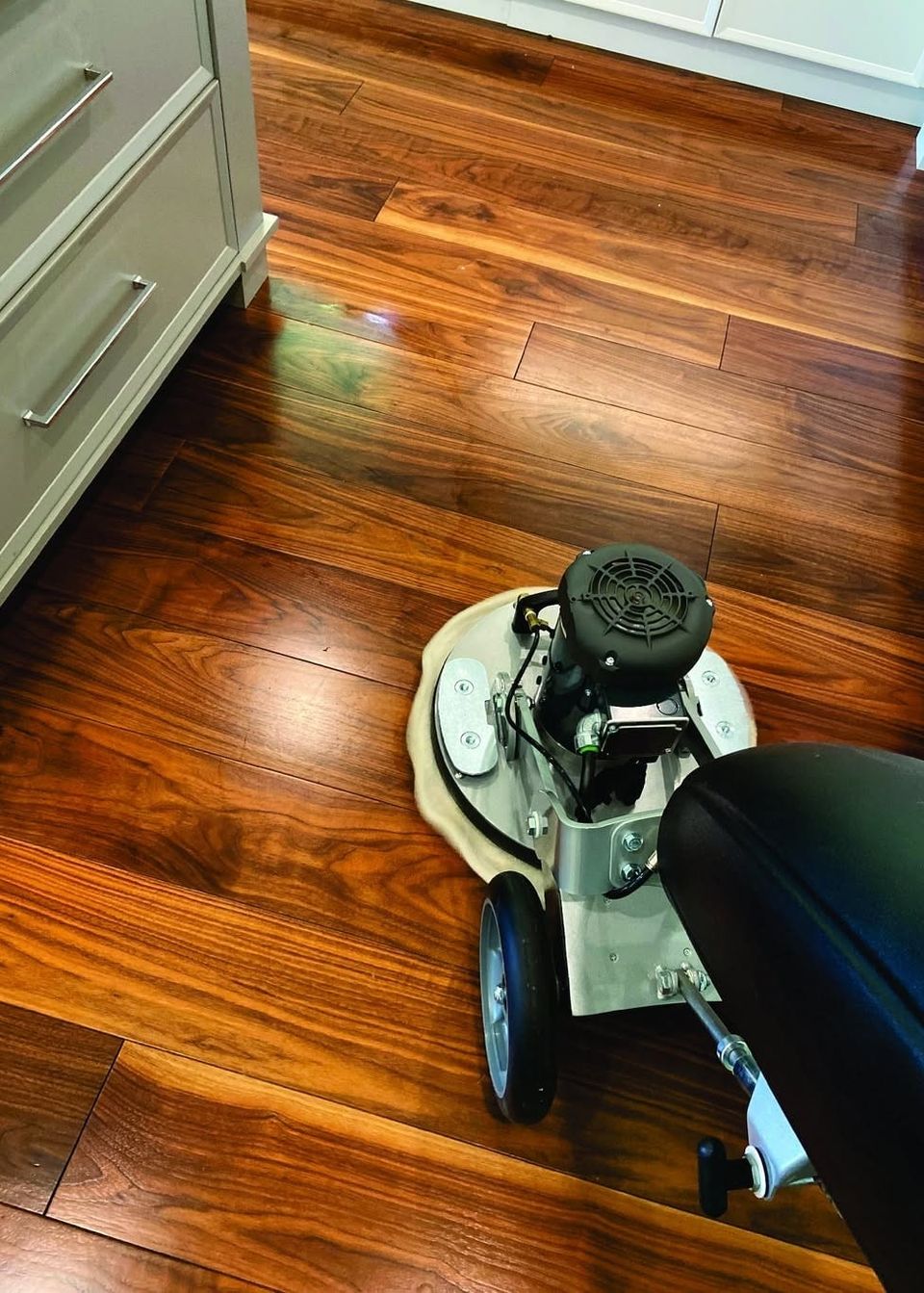 Mark's floor machine with a white cleaning pad on a glossy walnut hardwood floor beside kitchen cabinets